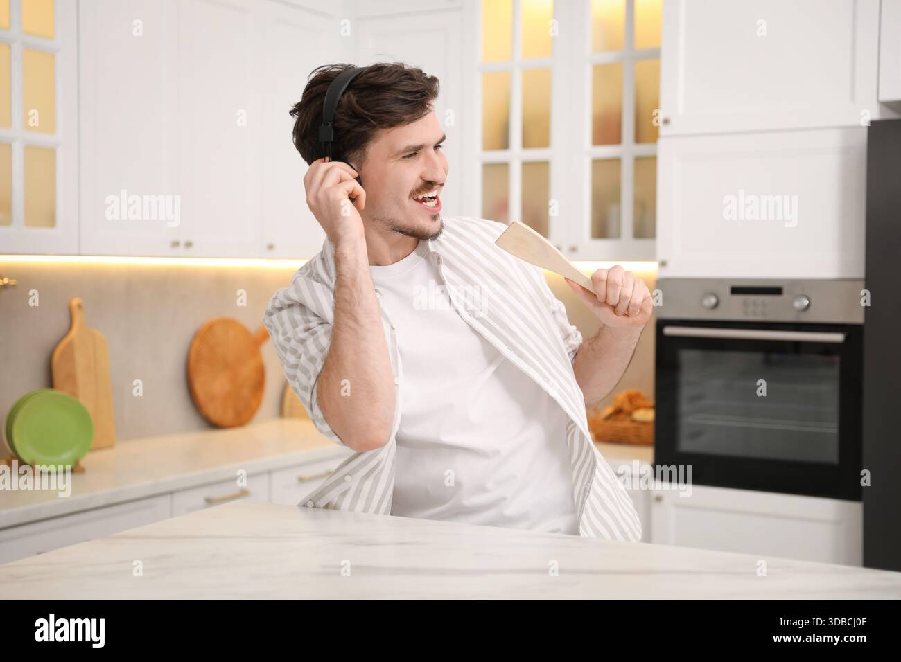 Happy man in headphones with spatula singing at white marble table in kitchen Stock Photo