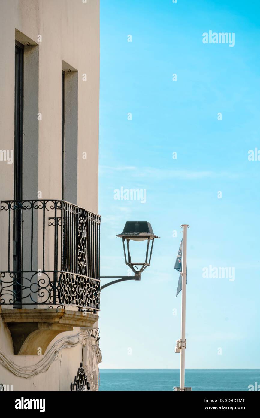 White stucco building with a wrought iron balcony, old street lamp, and flag overlooking the blue sea in Calella de Palafrugell, Spain Stock Photo