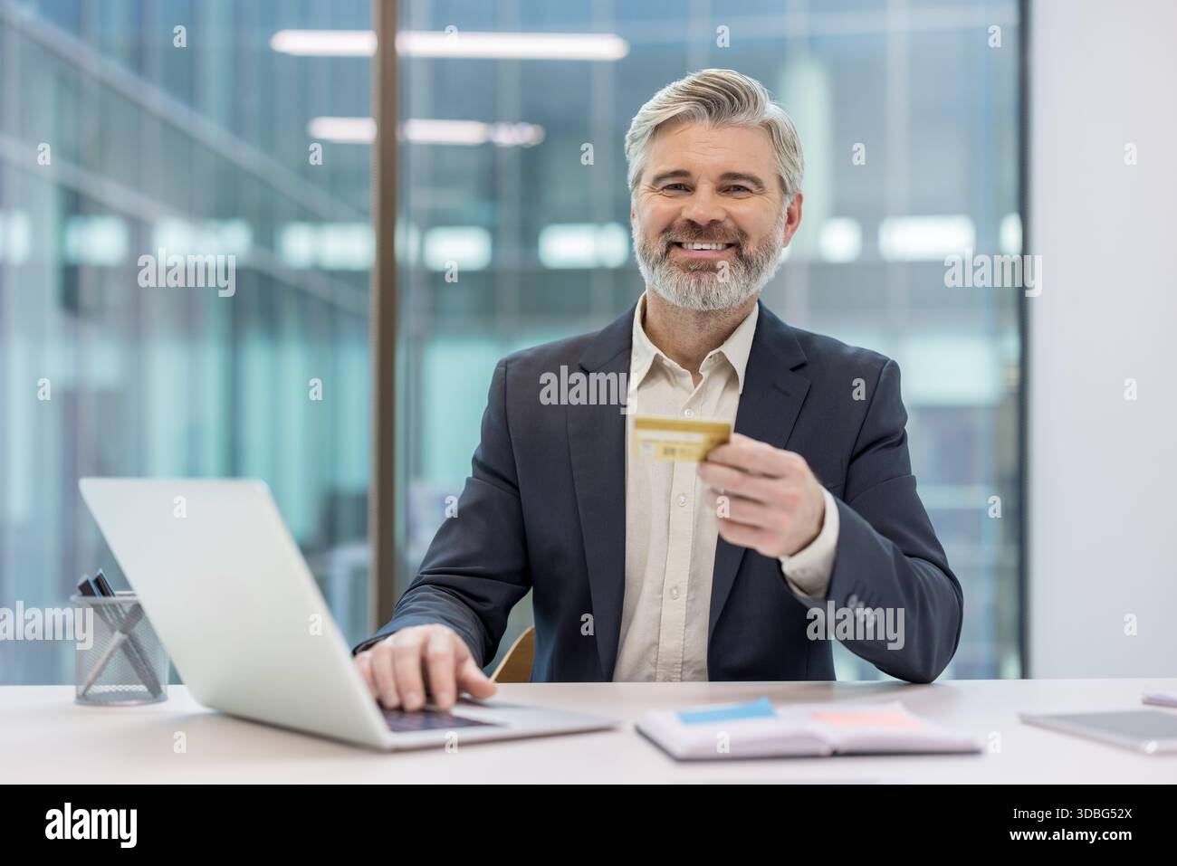 Mature businessman with a credit card smiling at the camera, using a laptop for online shopping and making secure e-commerce payments in a modern office setup Stock Photo