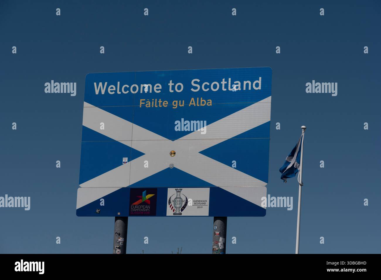 A blue road sign welcomes visitors to Scotland with Gaelic text and a nearby flag. Stock Photo