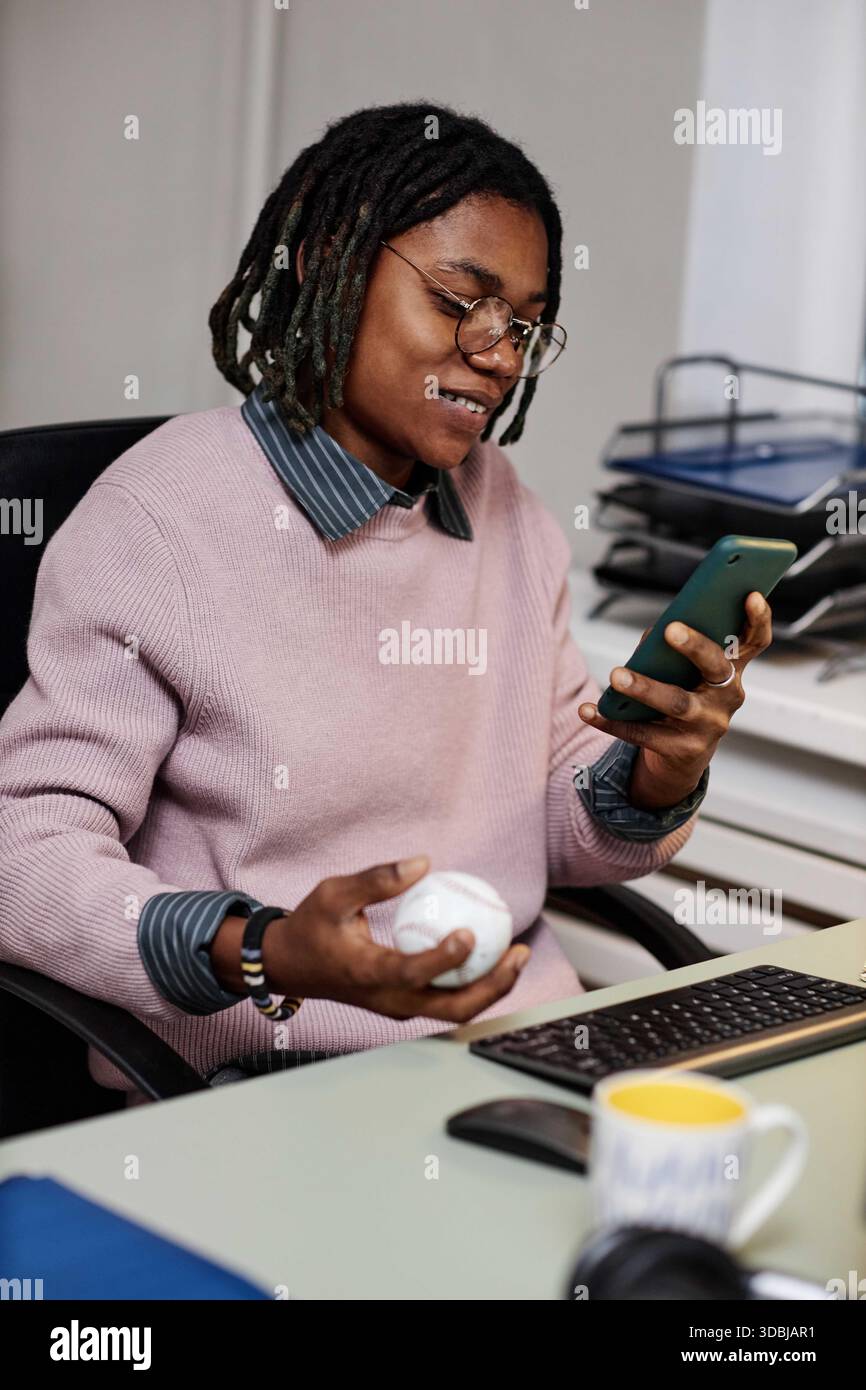 Young Adult Black Man Sitting at Desk Using Smartphone and Holding Baseball Stock Photo
