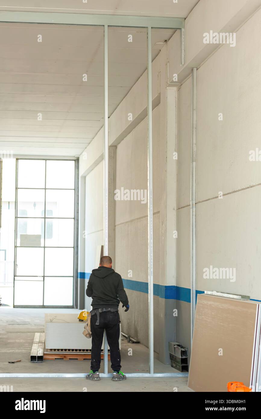 Construction worker installing aluminum pillars for the construction of a plasterboard wall in an industrial warehouse, using protective gloves and with his helmet hanging from his trousers Stock Photo