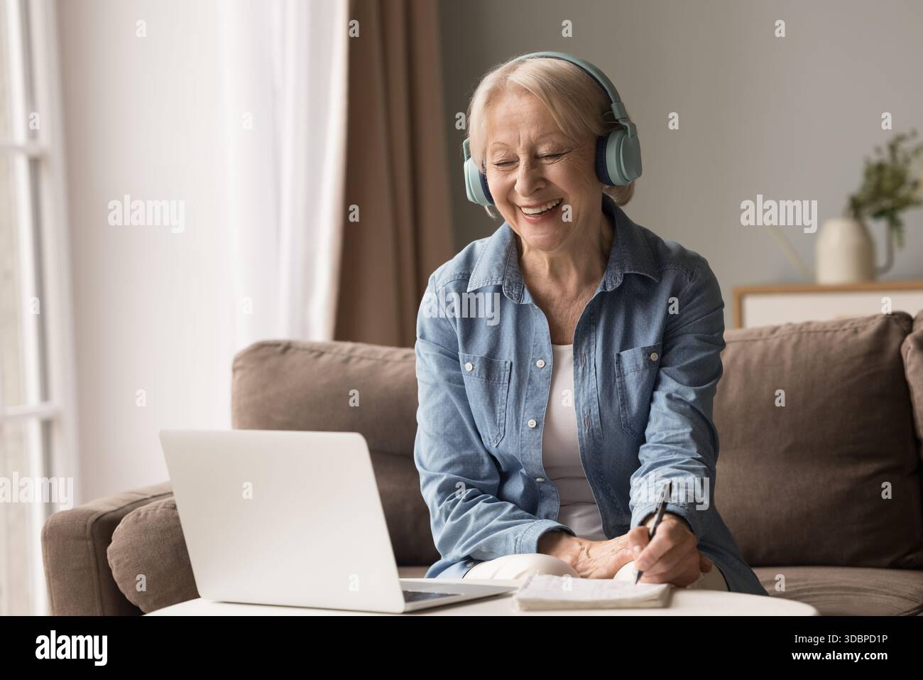 Aged lady in headphones learning online using laptop taking notes Stock Photo