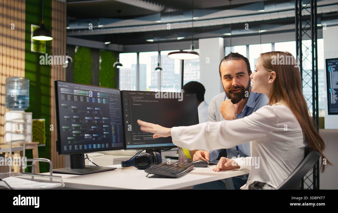 Friendly team leader helping happy young engineer code AI algorithms to debug software issues and improving system stability. Helpful manager assisting smiling intern in tech startup office, camera A Stock Photo