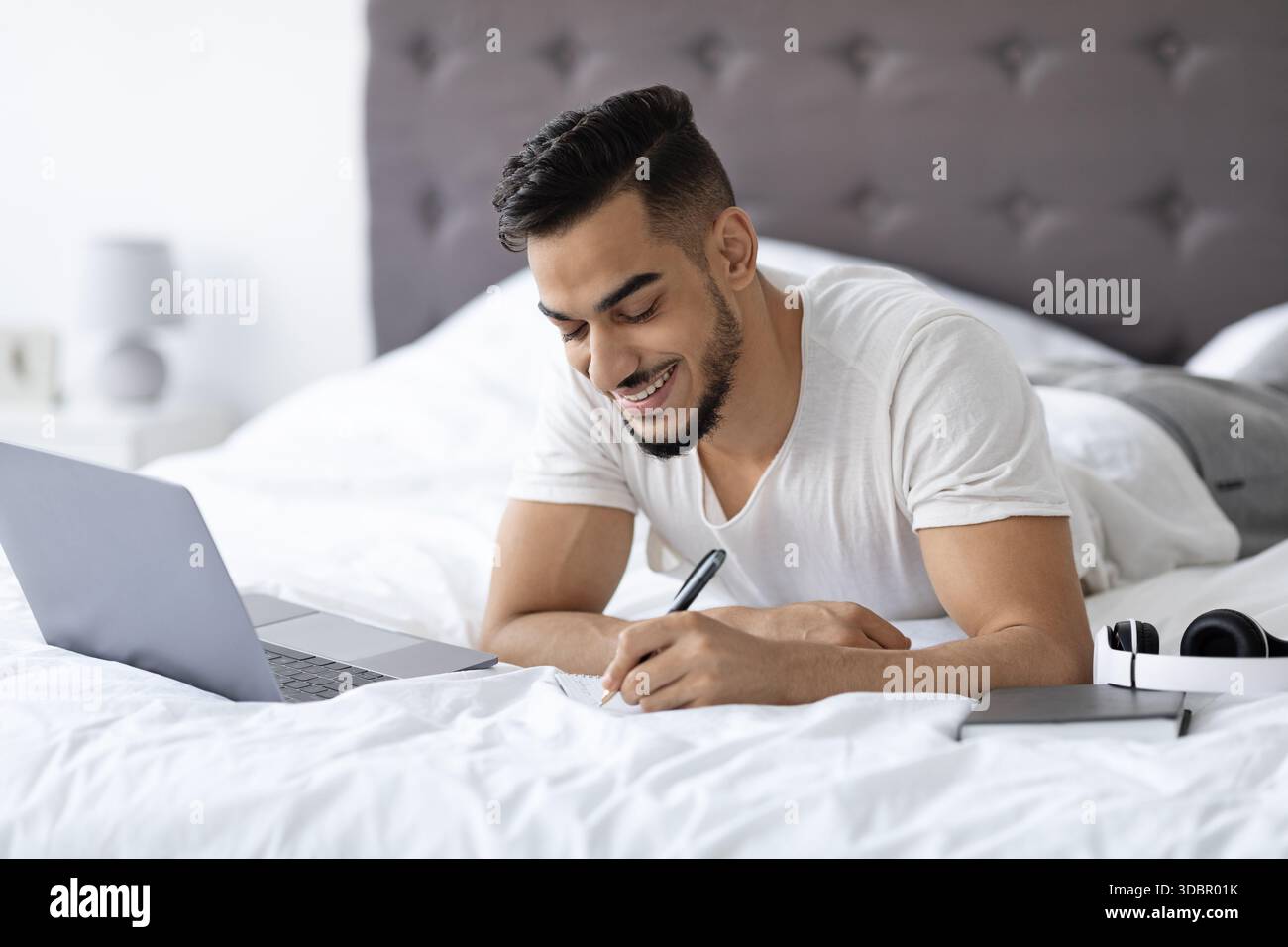 Young Arab Man Studies Online in Bed, Taking Notes on Distance Learning While Smiling Stock Photo
