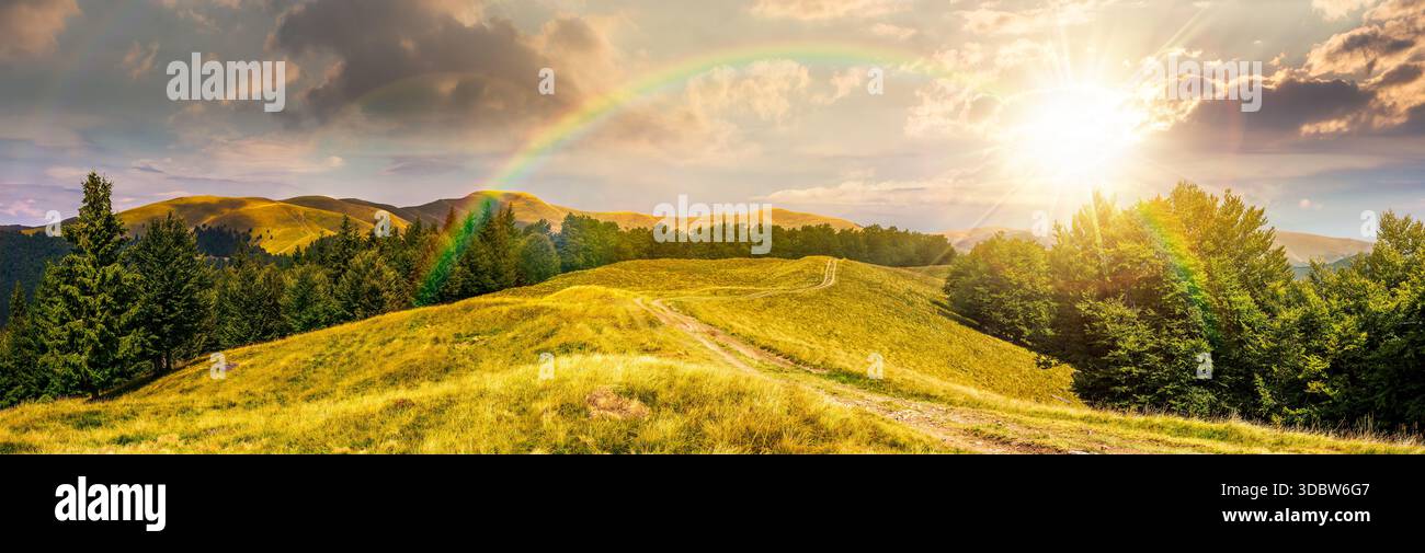 panorama of mountain ridge at sunset. country dirt road runs through grassy meadow near forest in evening light. location for hiking. beautiful weathe Stock Photo