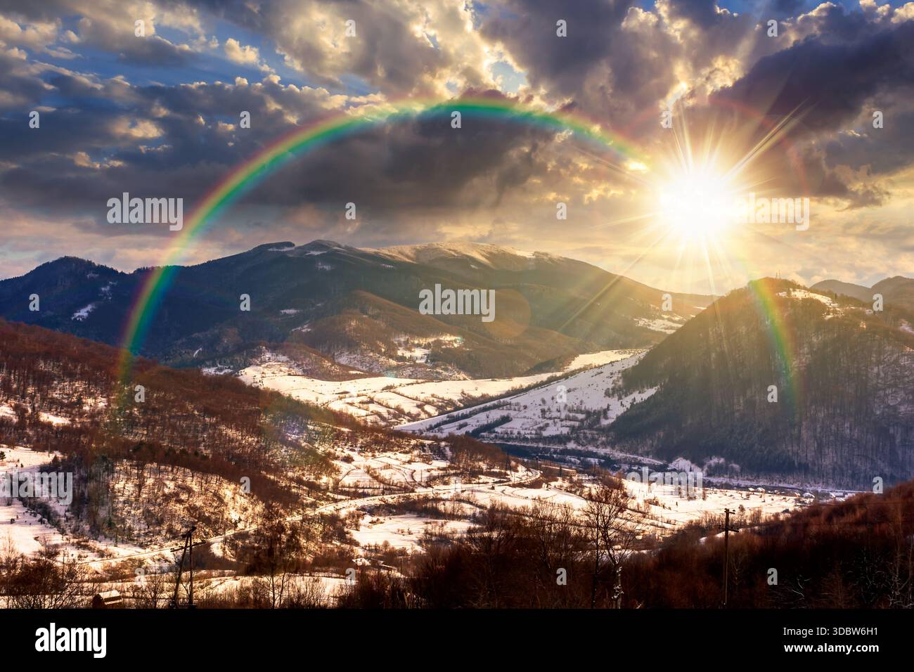 mountainous rural landscape in winter at sunset. snow covered fields on hills in evening light. village in the valley. landscape with clouds on sky. l Stock Photo