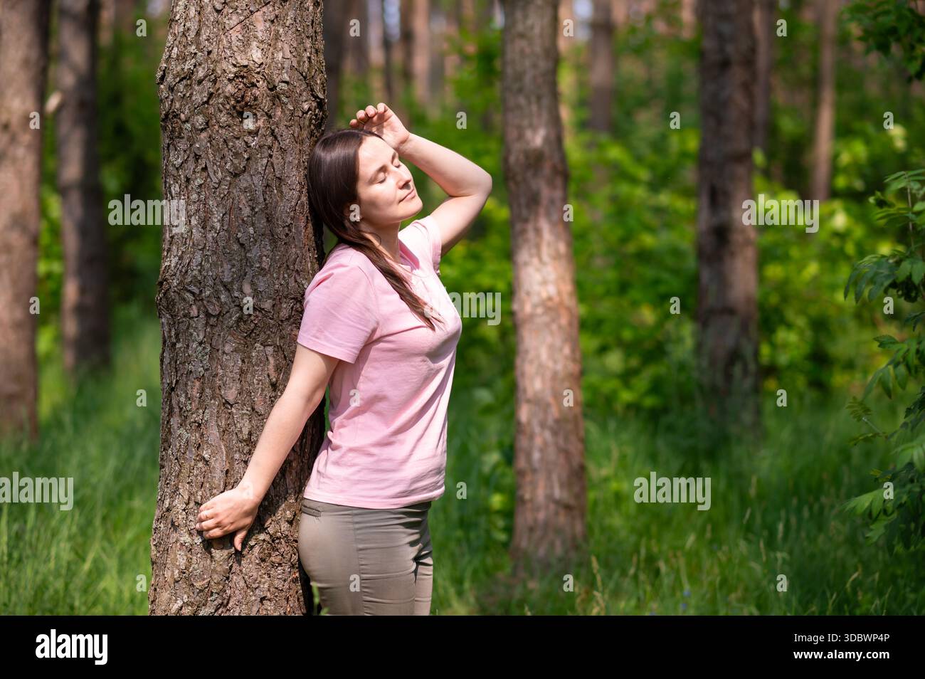 Young woman leaning against a tree in the forest, smiling and stretching her arms in the sun Stock Photo