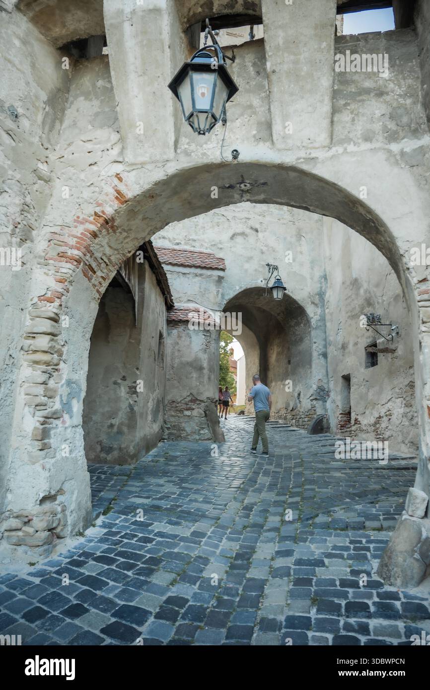 View of cobblestone street winding through aged stone archways, a lantern casts soft light on the medieval architecture, Sighisoara, Transilvania, Rom Stock Photo