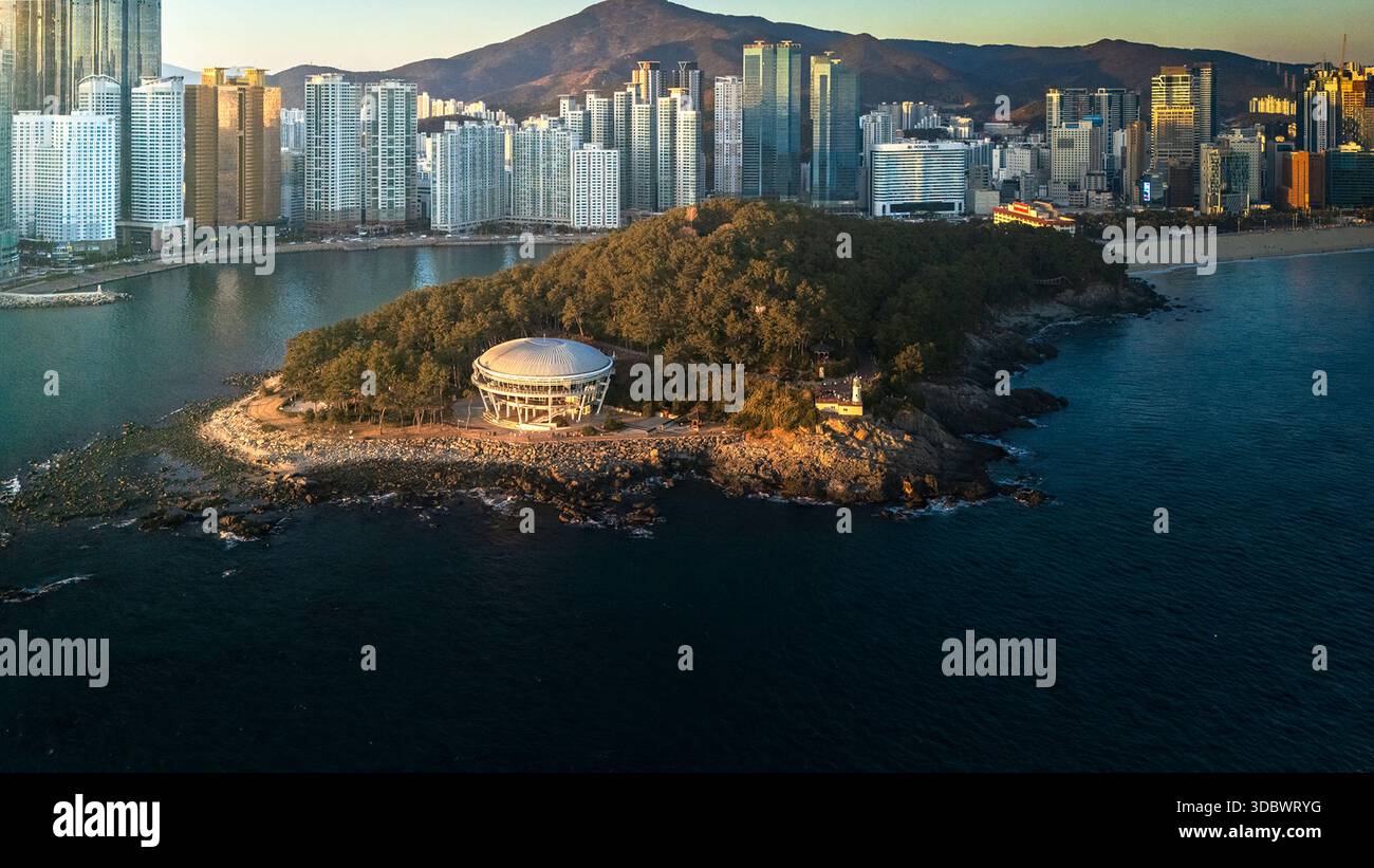 Aerial view of Dongbaek Island's lush greenery contrasts with the city skyline, reflecting the morning light on the water, Busan, Busan, South Korea. Stock Photo