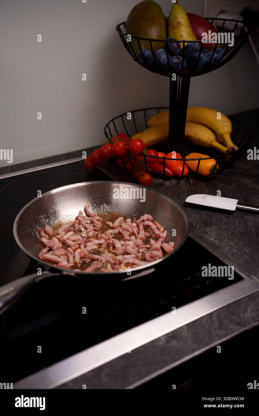 a stainless steel frying pan sizzles with bacon on an induction stove, flanked by a fruit stand laden with pears, mangos, bananas, tomatoes, and peppe Stock Photo