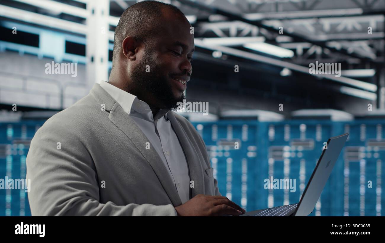 Data center engineer deploying AI driven management tools using laptop. Server farm employee inspecting server clusters, updating network topologies using artificial intelligence, camera B Stock Photo