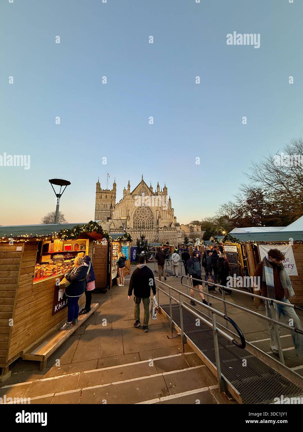 Exeter Cathedral Christmas Market at dusk, with visitors exploring festive stalls leading towards the cathedral’s west front. - Smartphone Captured Stock Image