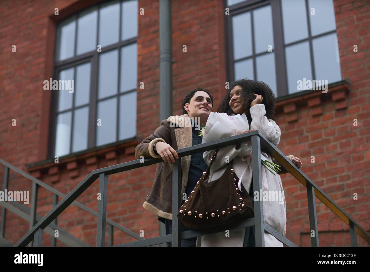 Young adult Caucasian man and young adult Black woman standing together on outdoor staircase, smiling and holding flowers, enjoying romantic moment in urban setting, dating context Stock Photo