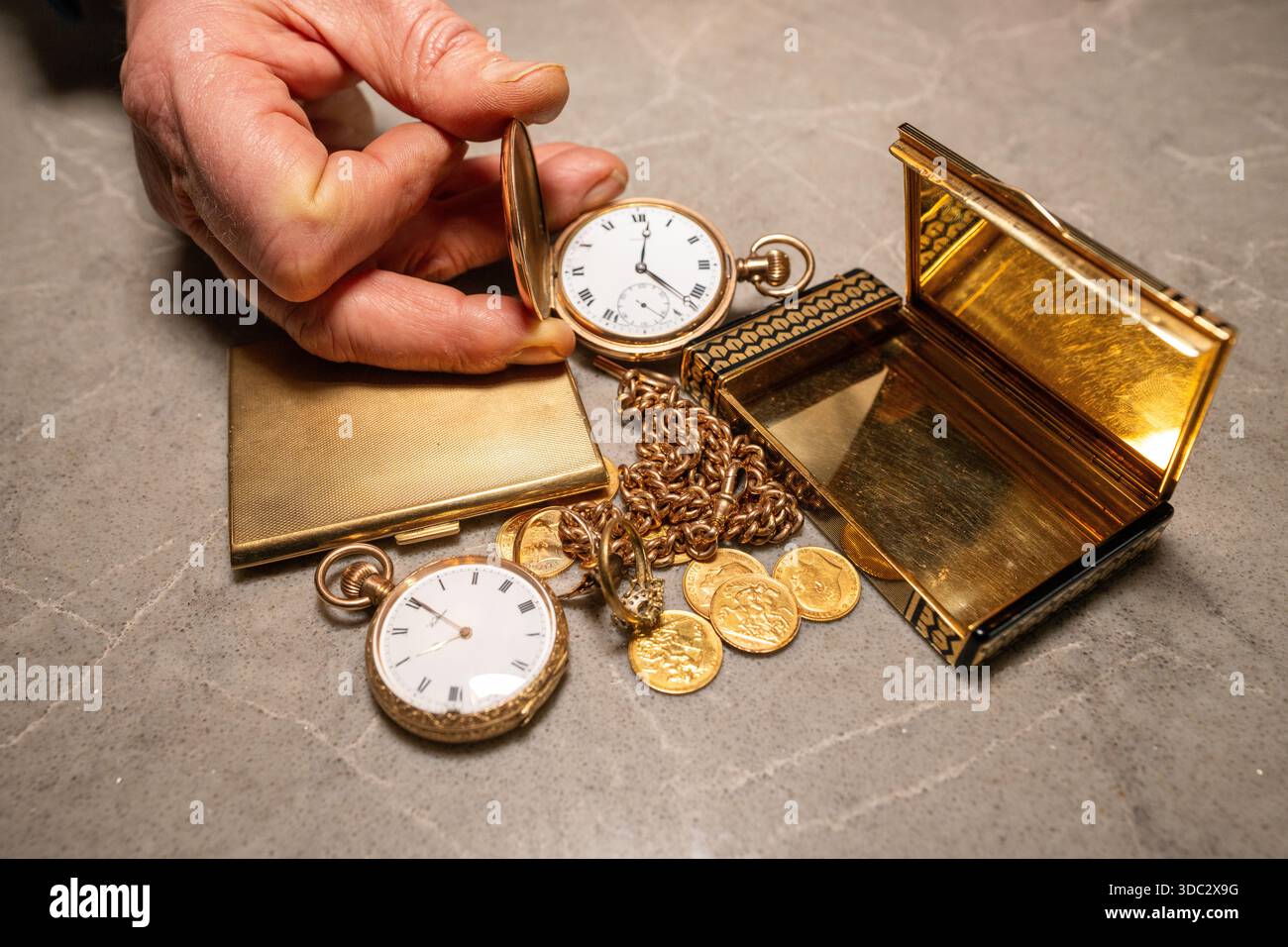 Man's hand holds old gold watch open with other gold items beneath Stock Photo