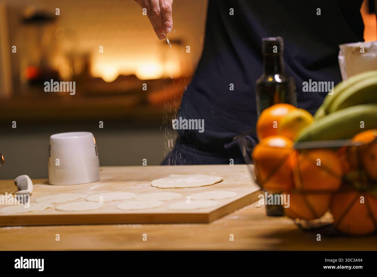 Rolled dough home kitchen cozy lifestyle moment. Unrecognizable person sprinkling flour above flattened pastry surface, warm evening light, calm domes Stock Photo