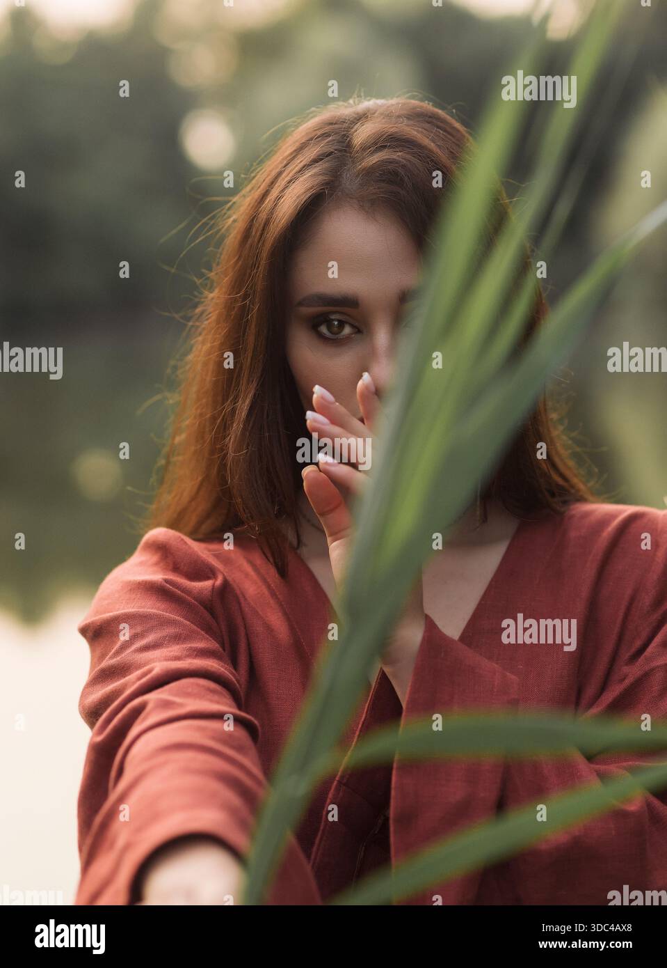 Cinematic shot of a woman in a reddish-brown robe sitting by a lake. The subject is framed by dense green reeds, looking directly forward. Soft lighti Stock Photo