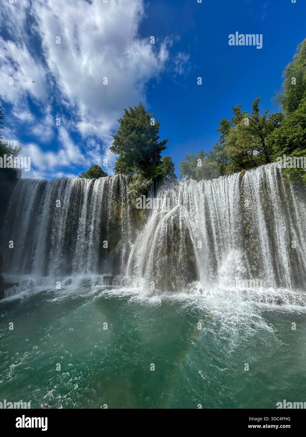 Wide waterfall cascading into a turquoise river, surrounded by lush green trees and dramatic clouds under a bright blue sky. Perfect for nature. - Smartphone Captured Stock Image