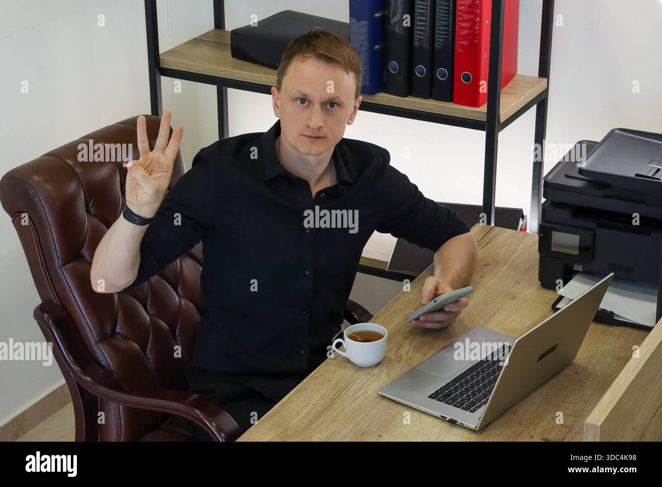 Professional at workstation shows three-finger gesture with one hand while using mobile device beside computer and tea. Stock Photo