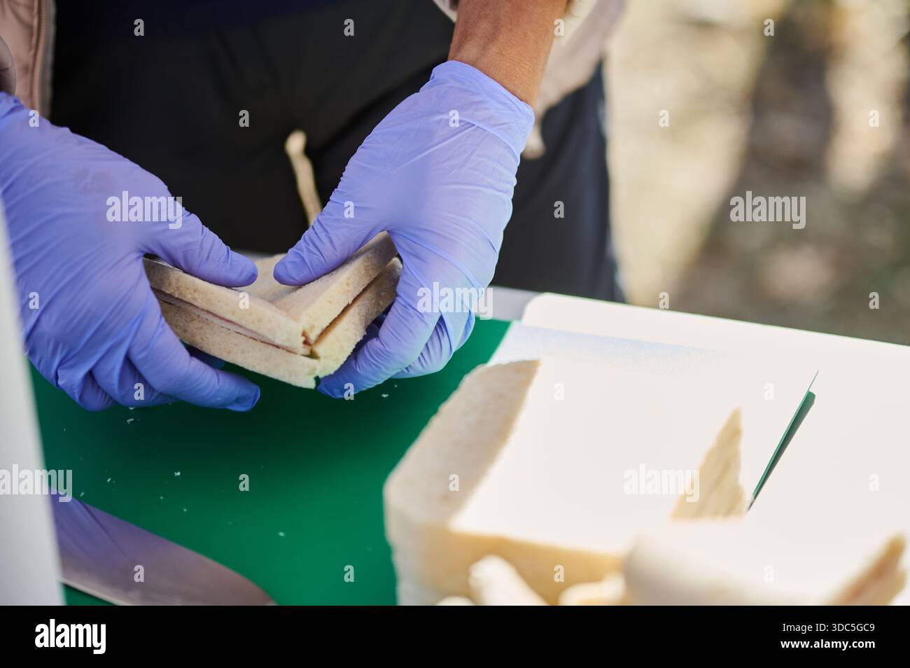 Gloved hands shape sandwiches on a green cutting board outdoors, with bread slices and a knife nearby, capturing casual food prep and outdoor cooking Stock Photo