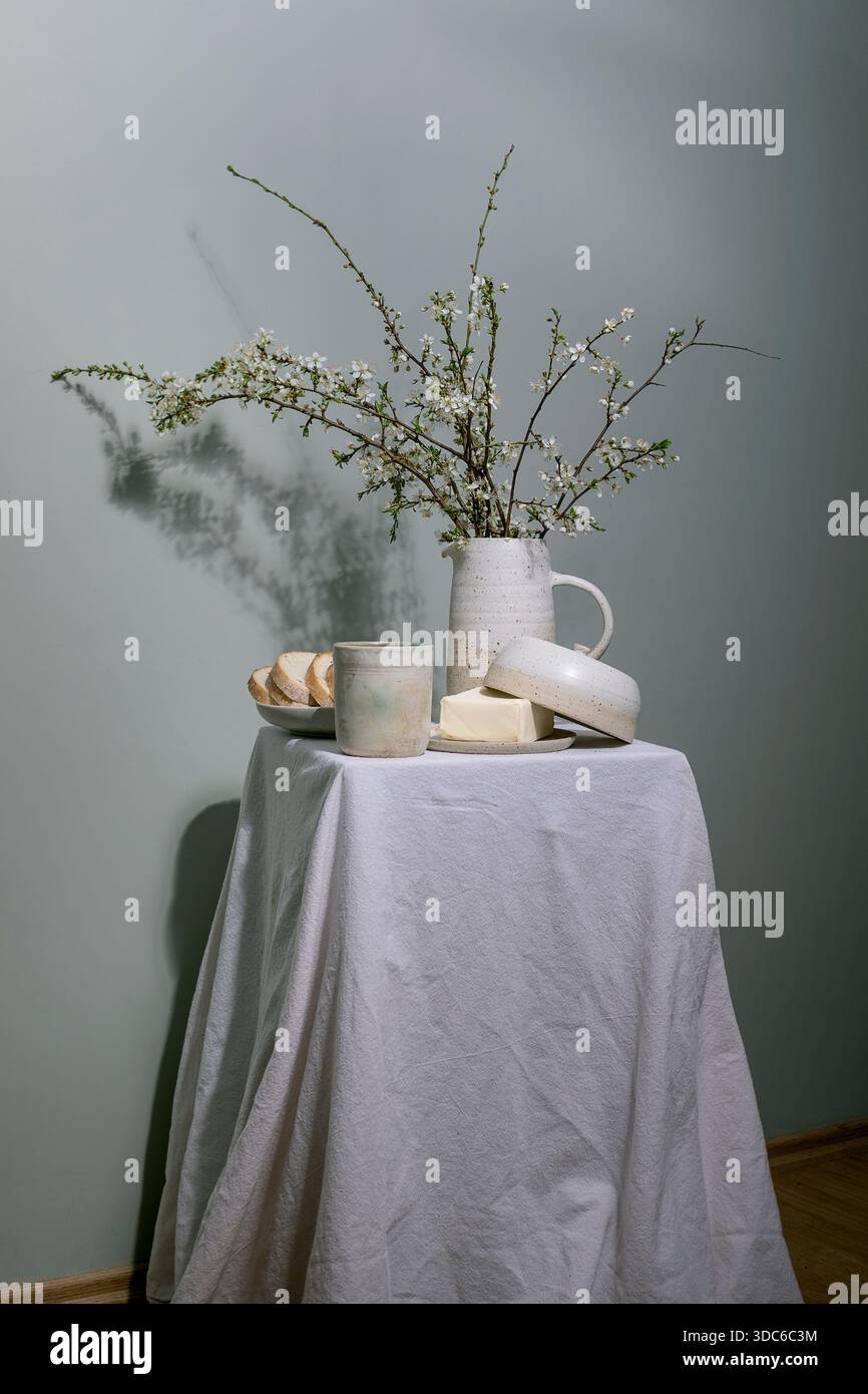 Minimal rustic breakfast setup with ceramic pitcher containing blooming cherry branches butter dish bread slice on plate ceramic cup on linen tableclo Stock Photo