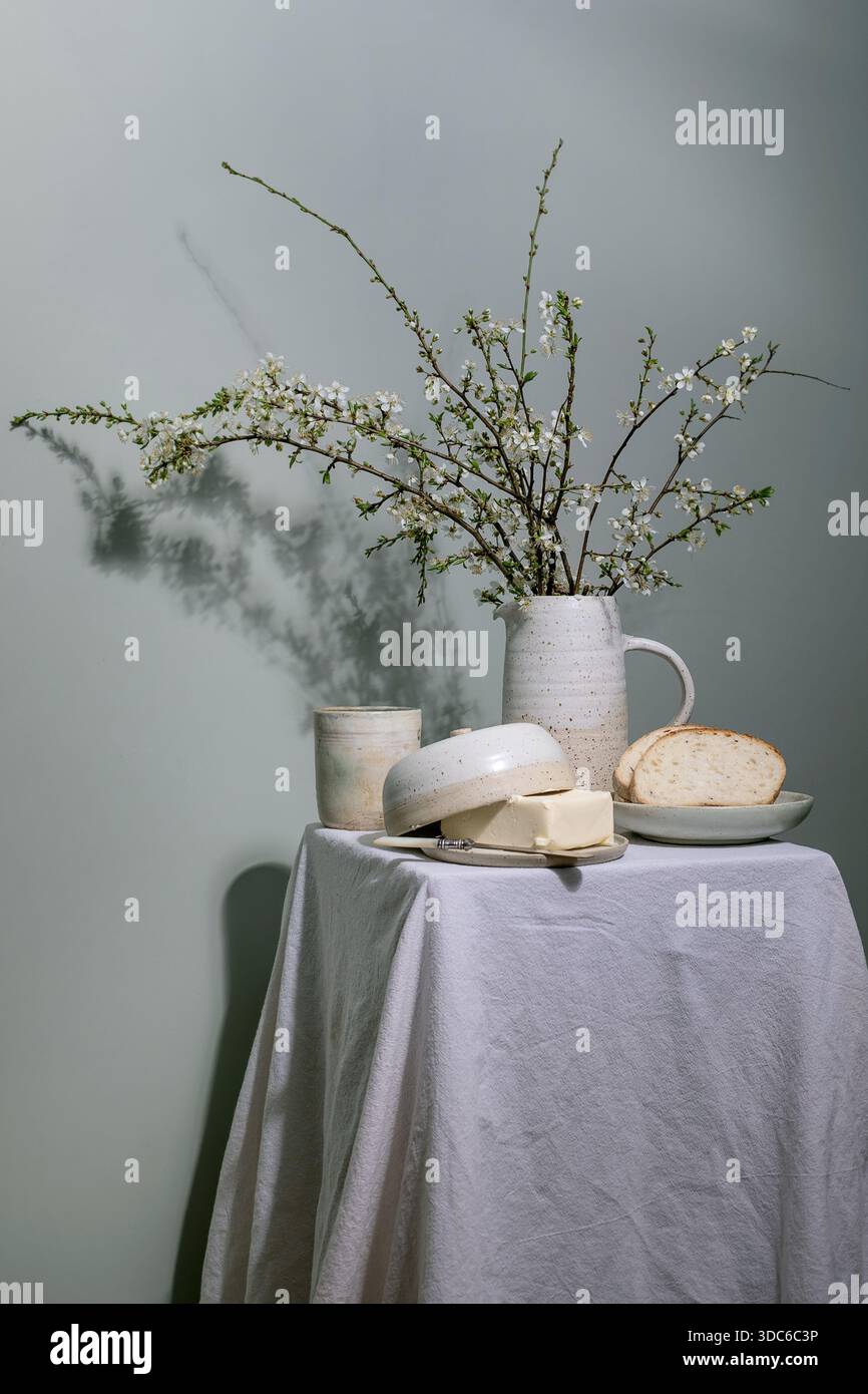 Minimal rustic breakfast setup with ceramic pitcher containing blooming cherry branches butter dish bread slice on plate ceramic cup on linen tableclo Stock Photo