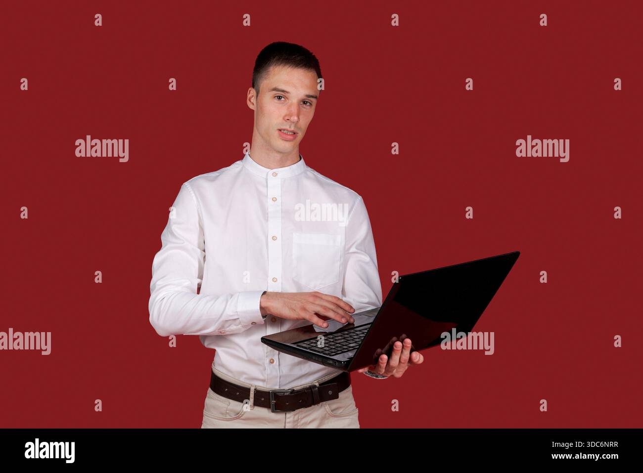 Portrait attractive focused cheerful brunette guy using laptop e-commerce project in white shirt isolated on bright red color background || Model released Stock Photo