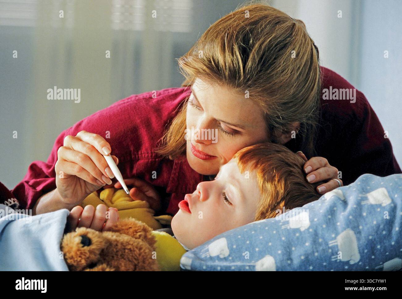 A mother comforts her sick child in bed. She uses a thermometer to check his temperature. The child looks tired and is lying under a blanket with a te Stock Photo