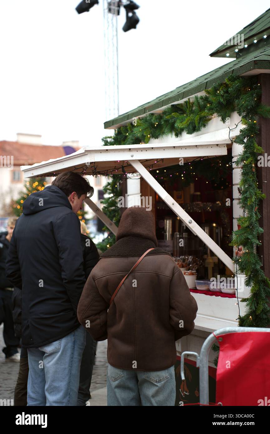 people in winter attire visit a festive market stall decorated with greenery and lights, set against a urban backdrop with cobblestone path and christ Stock Photo