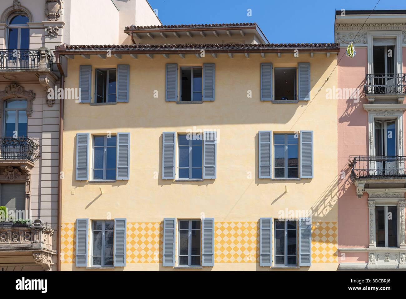 Sunny European street scene featuring a warm yellow facade, blue shutters,  and decorative balconies. Bright, colorful architecture with tiled lower wa  Stock Photo - Alamy, image size:1300x956