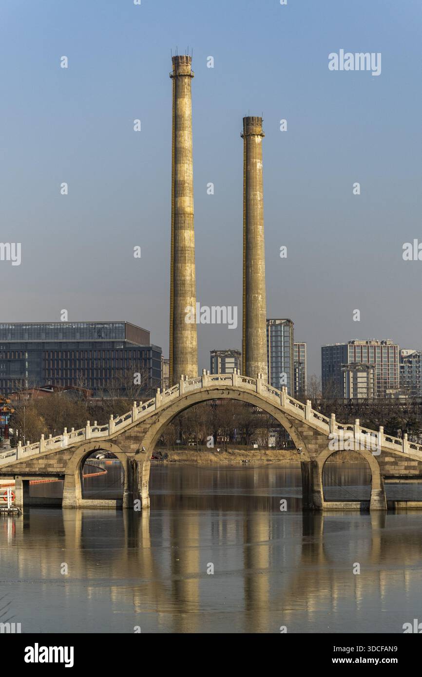 View of a serene bridge reflecting on the water against the backdrop of towering industrial chimneys under a clear sky, Beijing, Beijing, China. Stock Photo