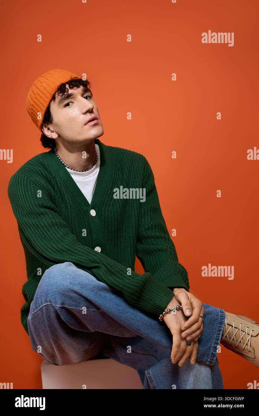 A handsome man in stylish attire sits confidently against a bright orange wall. Stock Photo