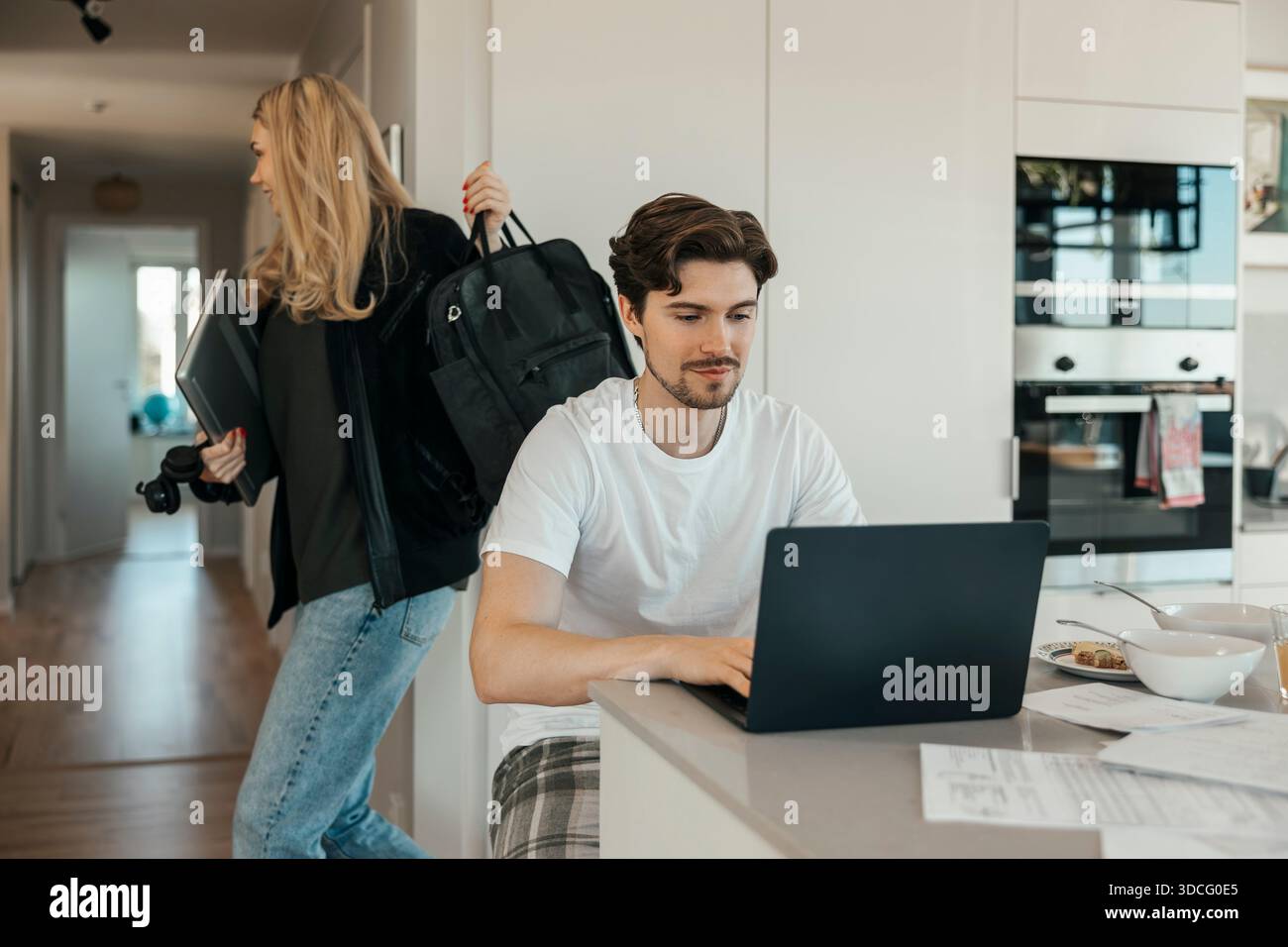 Young man using laptop at home while girlfriend leaving for work Stock Photo