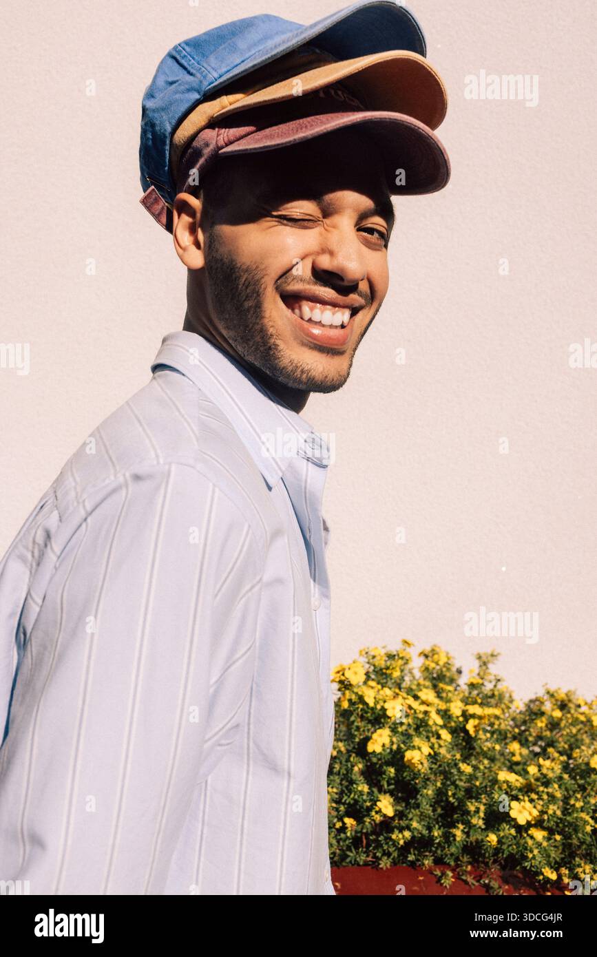 Smiling man winking while wearing multi colored caps against wall Stock Photo