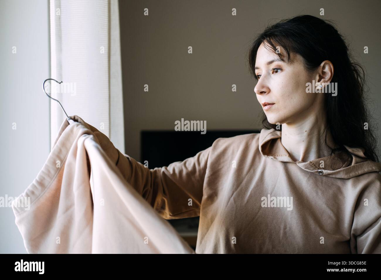 Woman looks at garment on hanger near window. High-quality basics, slow fashion movement, textile sustainability. Stock Photo
