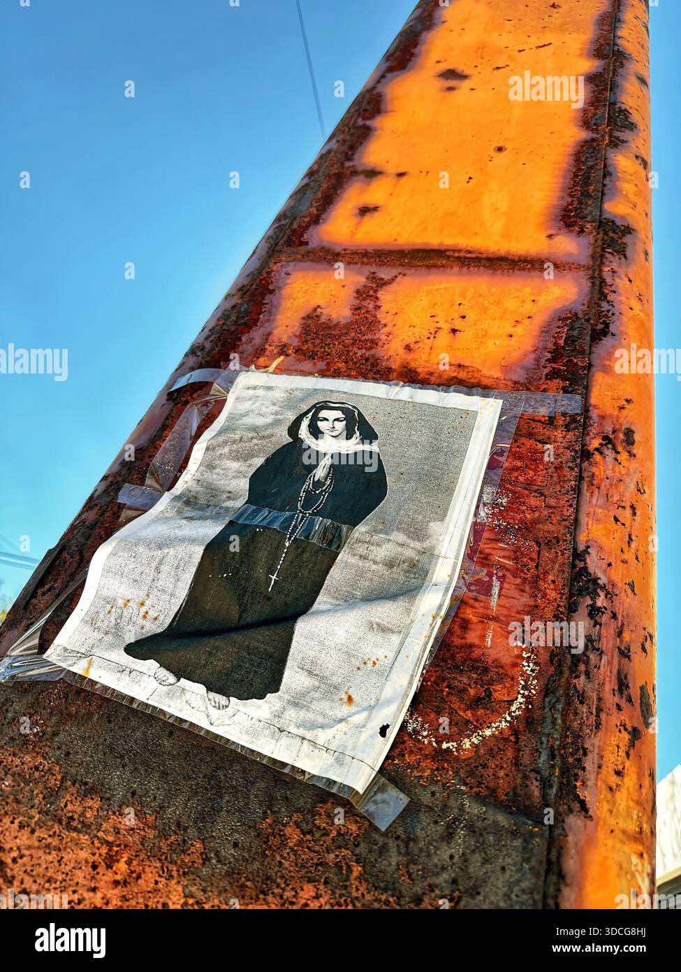Poster of a hooded figure with a rosary on a weathered rusted metal pole under a clear blue sky in an urban outdoor setting Stock Photo