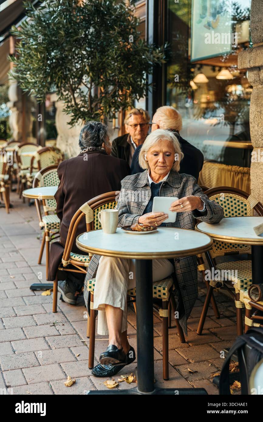 Senior woman using tablet PC while sitting at table in sidewalk cafe Stock Photo