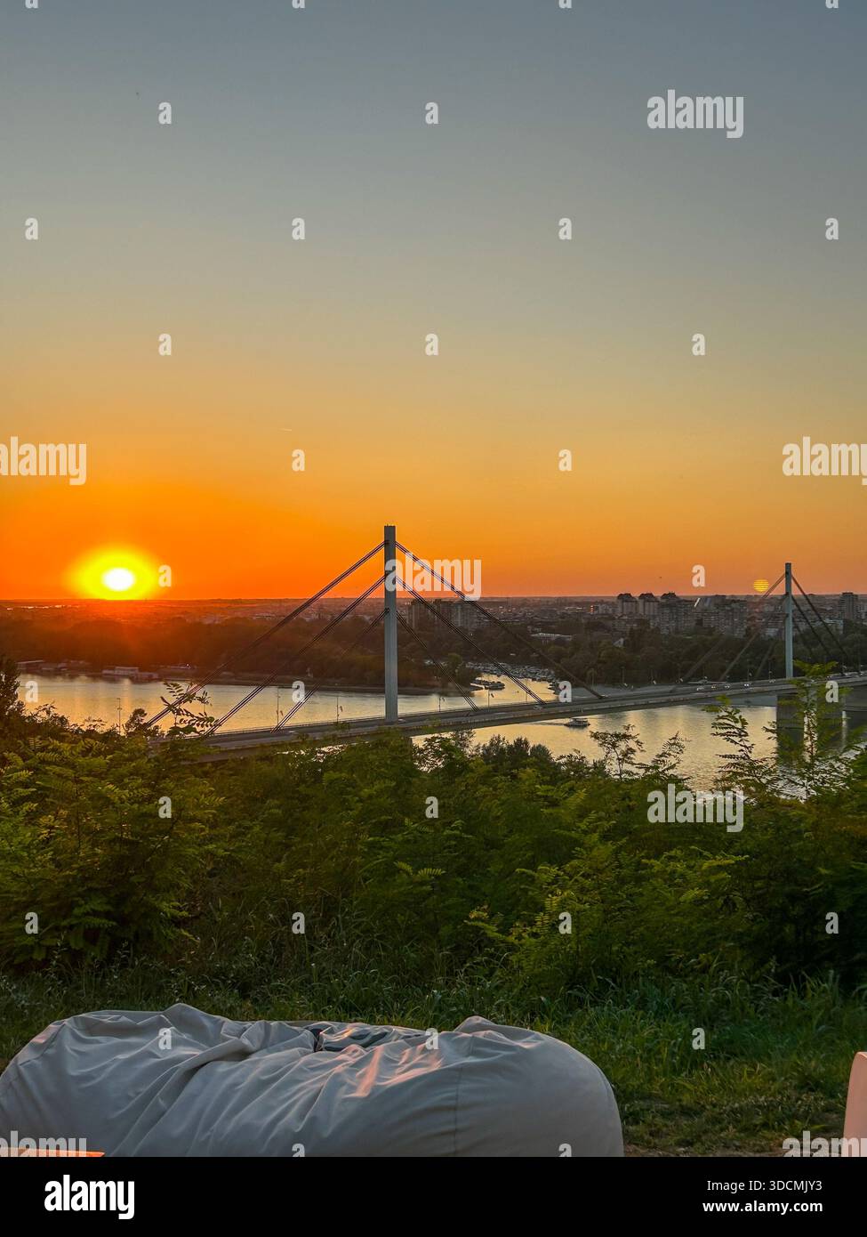 Warm sunset over the Liberty Bridge spanning the Danube River in Novi Sad, with city skyline and reflections creating a peaceful atmosphere. - Smartphone Captured Stock Image