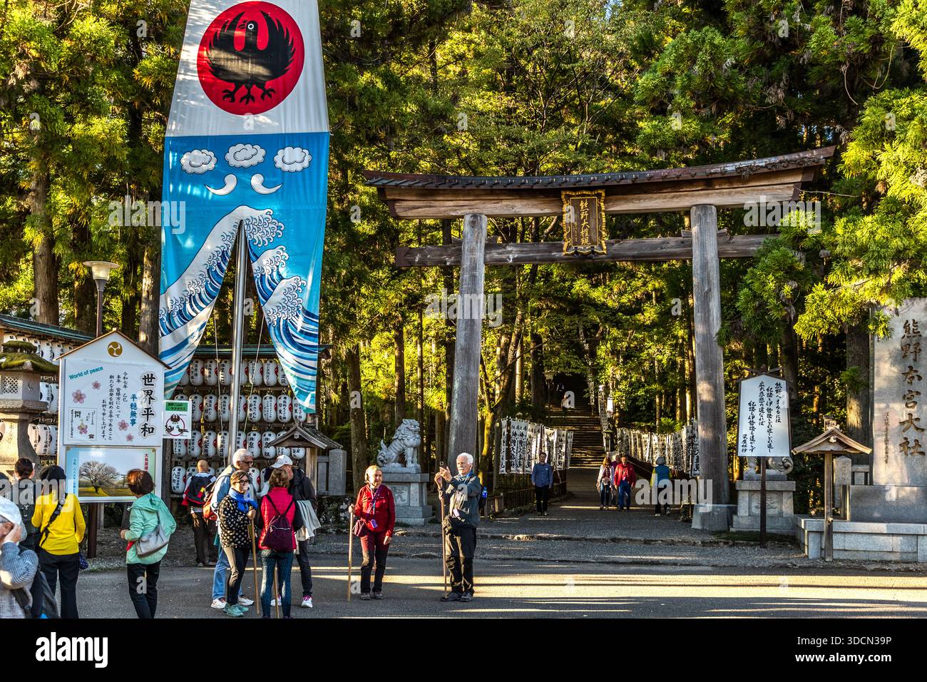 Visitors photograph a flag featuring Yatagarasu, the three-legged crow, in front of a torii at the Oyunohara Approach to Kumano Hongū Taisha, Tanabe, Wakayama Prefecture, Japan. Yatagarasu is regarded as the pilgrims' guide and a symbol of the Kumano Kodo. Stock Photo