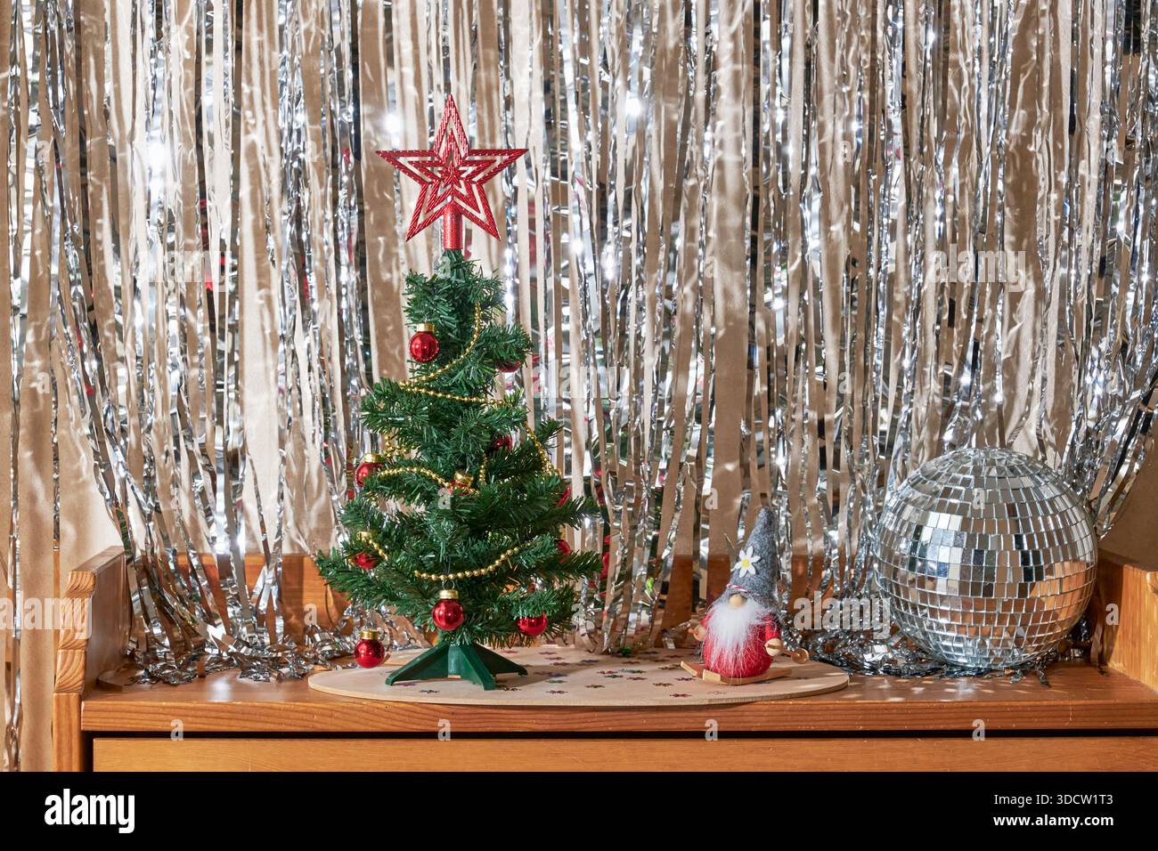 Cheerful Christmas composition. Small decorated fir tree with red star topper, shiny disco ball, and Nordic gnome on skis figurine on wooden surface w Stock Photo