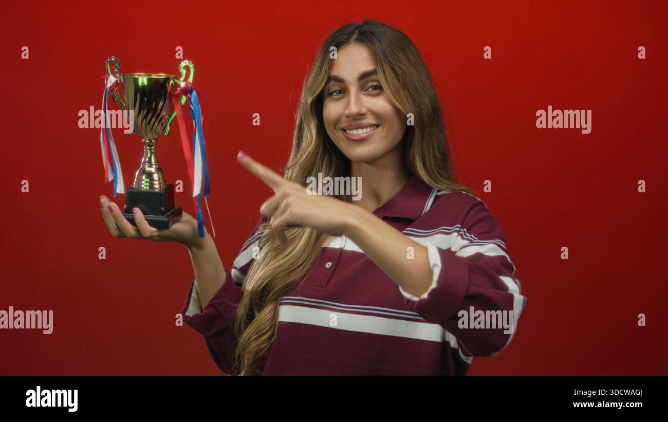 Woman holds trophy in studio with finger pointing to trophy highlighting the cup against red background; achievement celebration success pride. Stock Photo