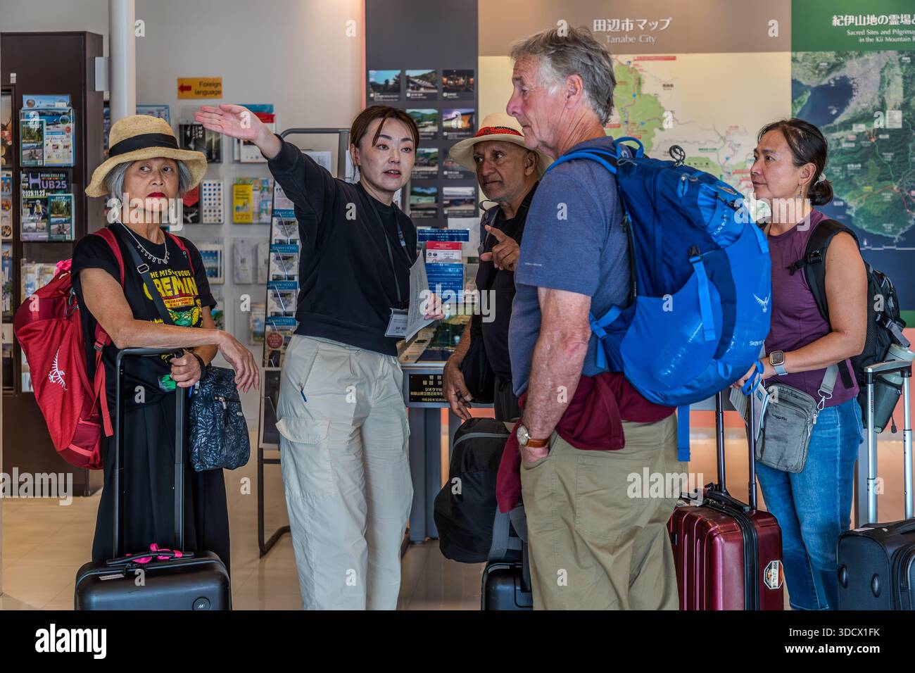 Travellers with suitcases receive advice from a staff member at the tourist information centre in Tanabe, Wakayama Prefecture, Japan, who points to a map of the Kii mountains. Tanabe is the starting point for a popular stage of the Kumano Kodo, a historic network of pilgrimage routes on the Kii Peninsula that has been a UNESCO World Heritage Site since 2004 and is often called the "Japanese Way of St James". Stock Photo