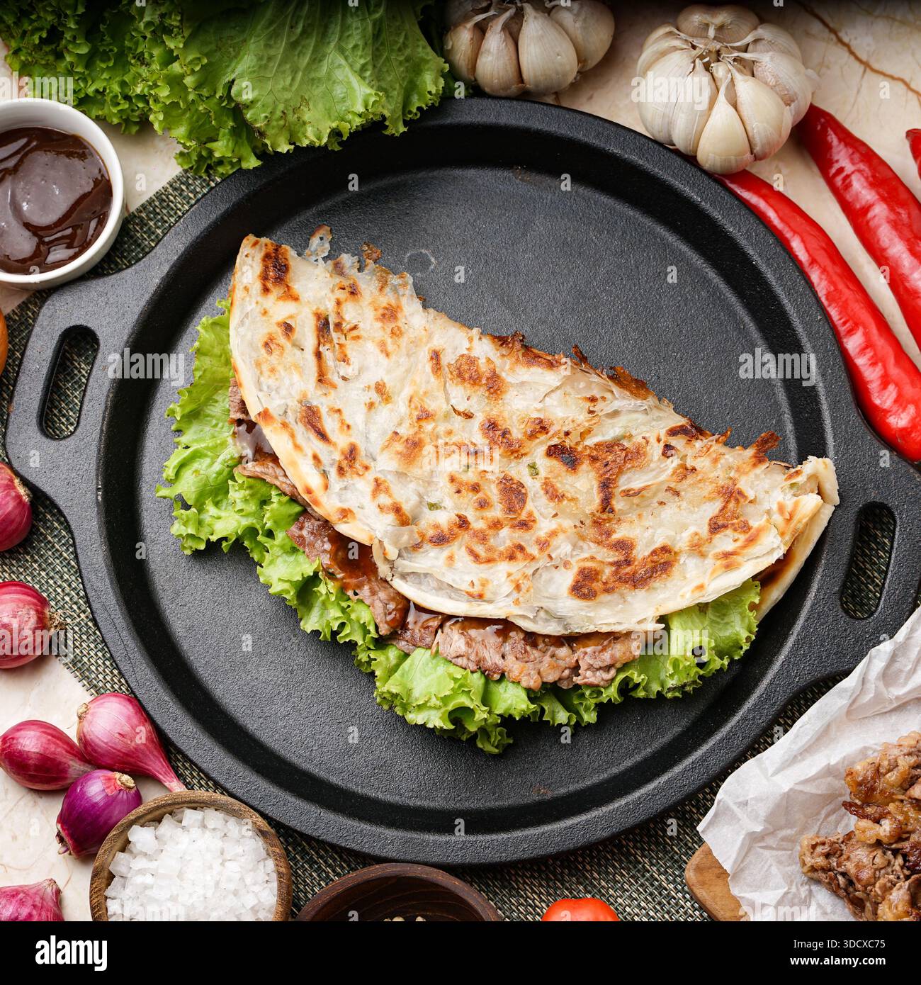 Savory quesadilla filled with meat and lettuce, served on a cast iron skillet surrounded by fresh ingredients like red onions, garlic, and chili peppe Stock Photo