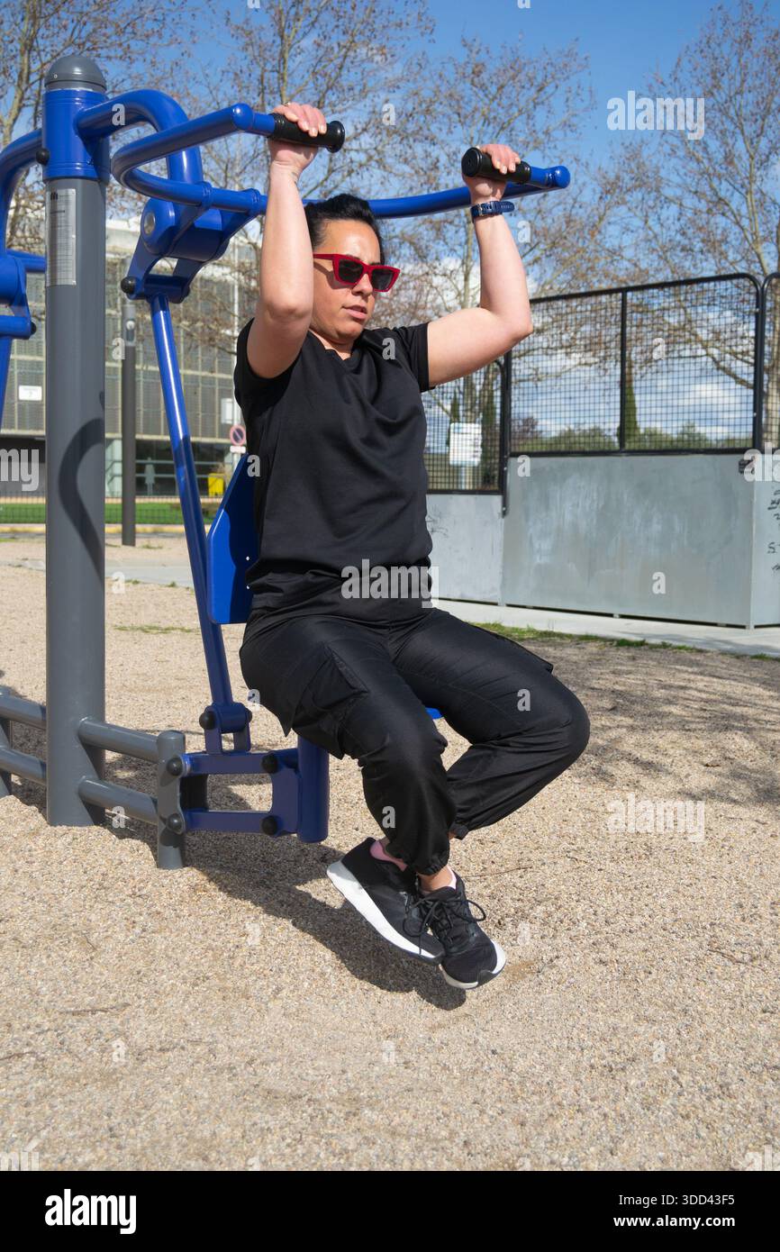 Woman performing pull-ups on an outdoor gym machine, enjoying a sunny day workout in the park Stock Photo