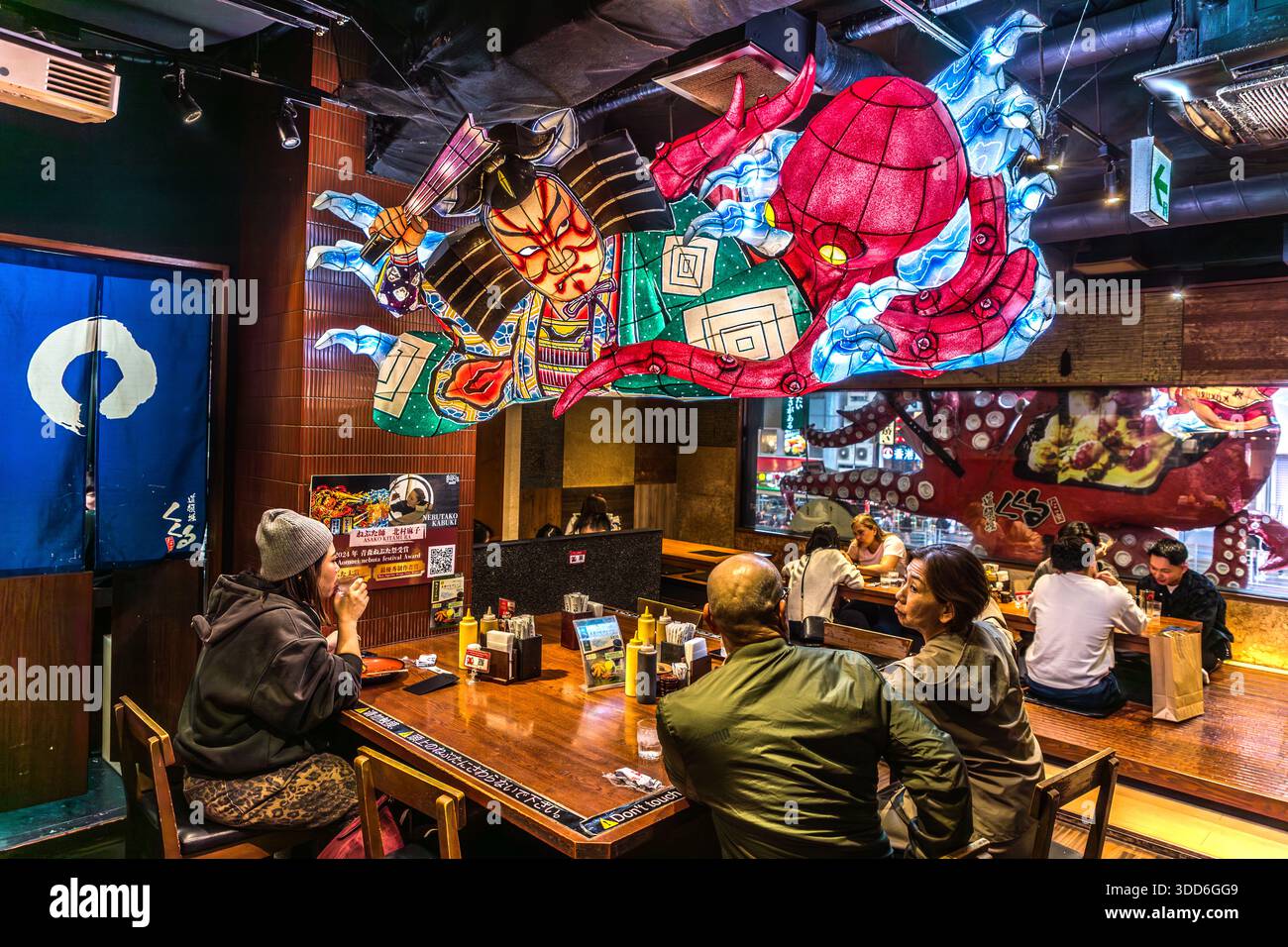 Guests sit in a takoyaki restaurant in Osaka's Dōtonbori entertainment district, Japan, beneath a colourful ceiling decoration of a samurai mask and an octopus. Takoyaki, fried octopus balls, are considered the city's signature dish and are served both as street food and in restaurants. Street vendor Tomekichi Endo invented the dish in the 1930s. Stock Photo