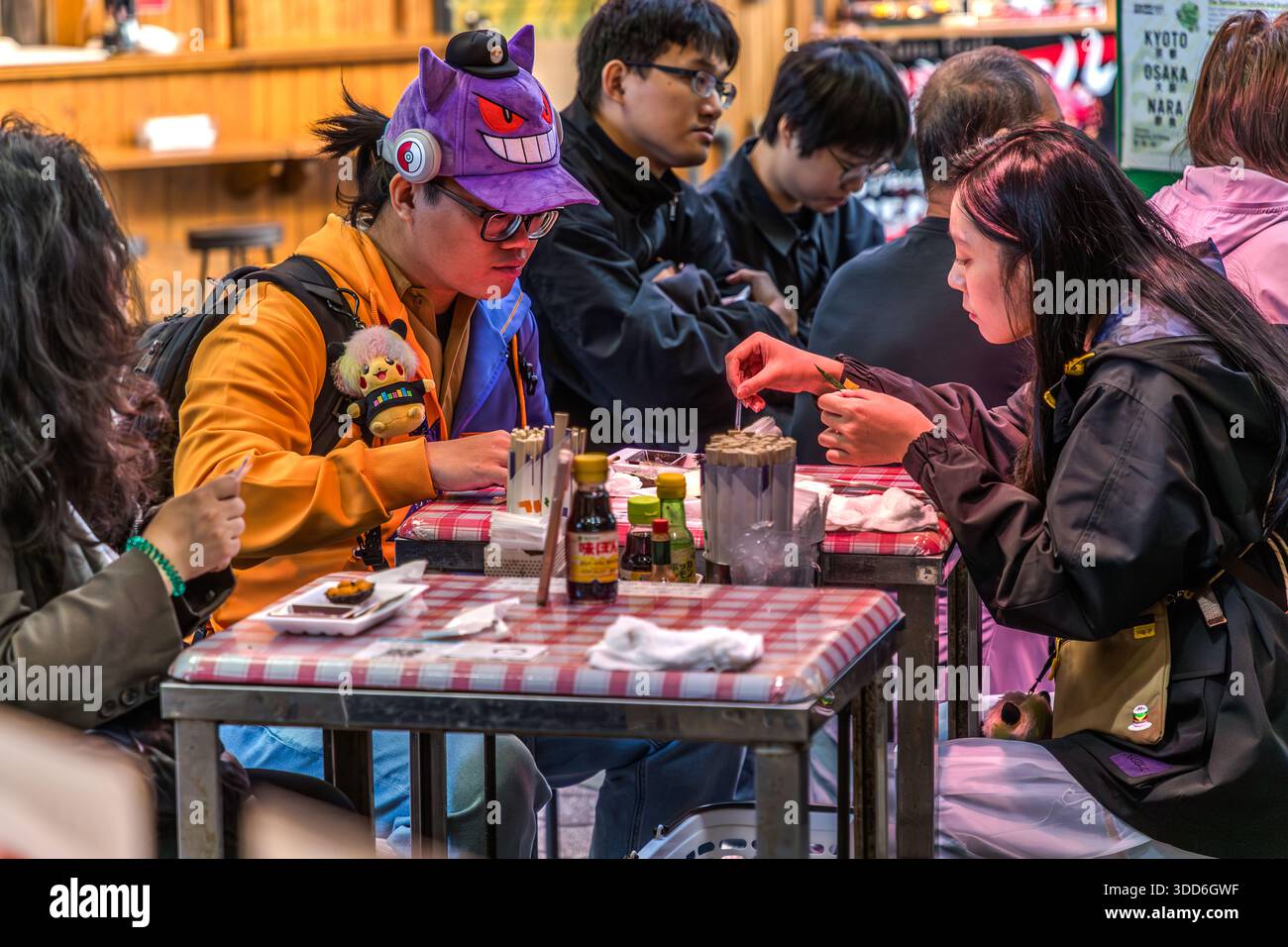 A smiling cap on your head and a Pokémon on your rucksack: Nuigurumi Charms is the name of the trend where all kinds of soft toys are worn on rucksacks, clothing or as caps. Young customers with fan merchandise accessories from otaku or gaming scenes in a market restaurant in Osaka, Osaka Prefecture, Japan Stock Photo