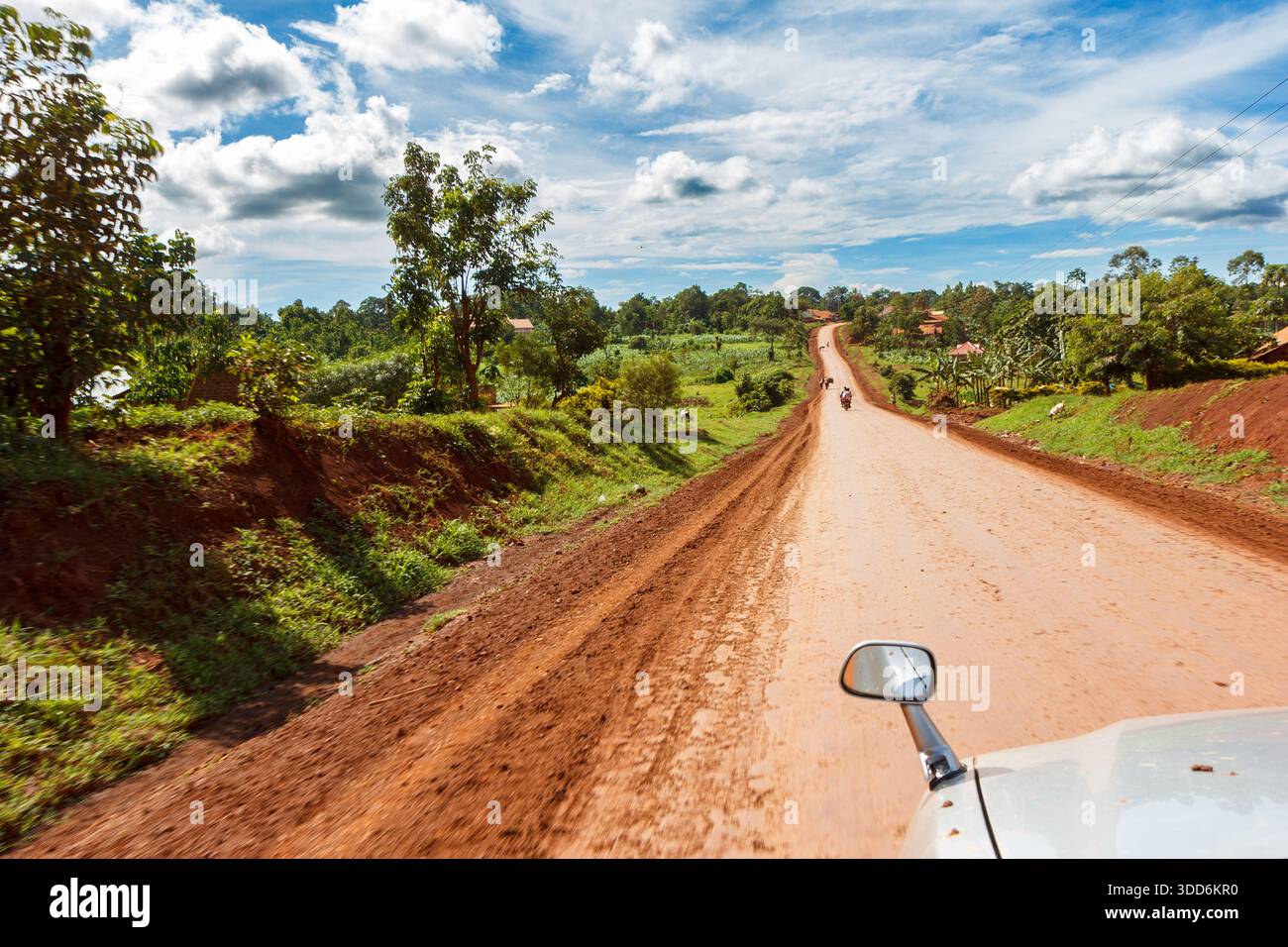 A reddish dirt road in a Ugandan rural area, surrounded by lush greenery. - Stock Image
