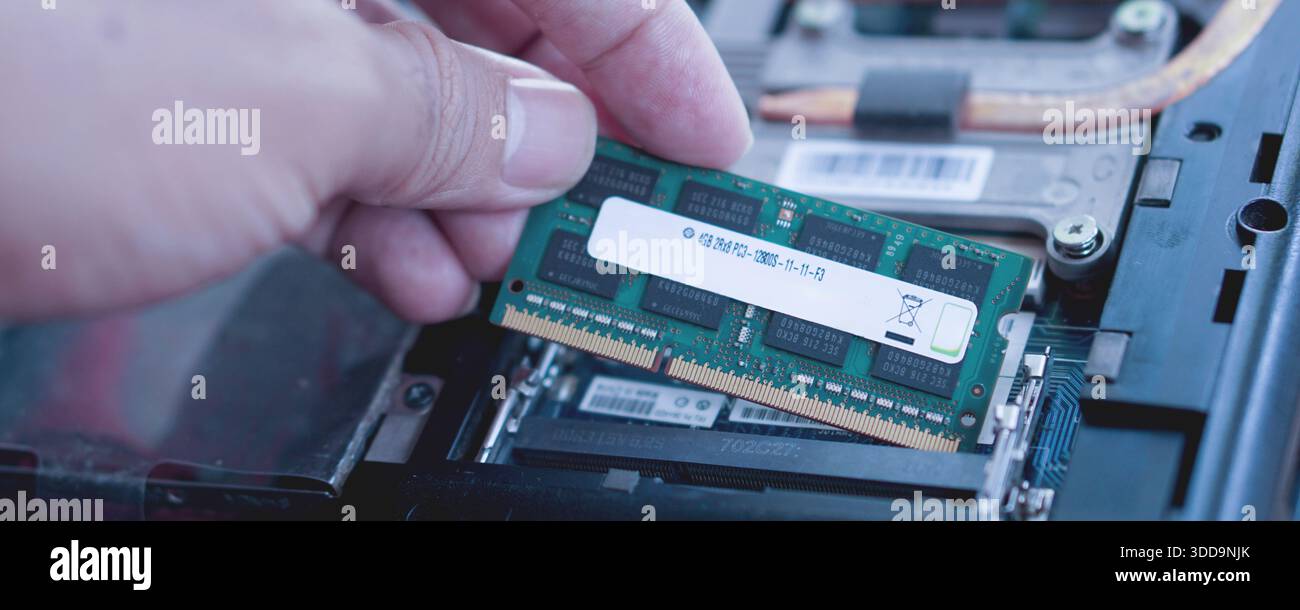 A technician's hand carefully inserting a green RAM stick into the laptop's memory slot. Visualizes hardware maintenance and performance tuning. Stock Photo
