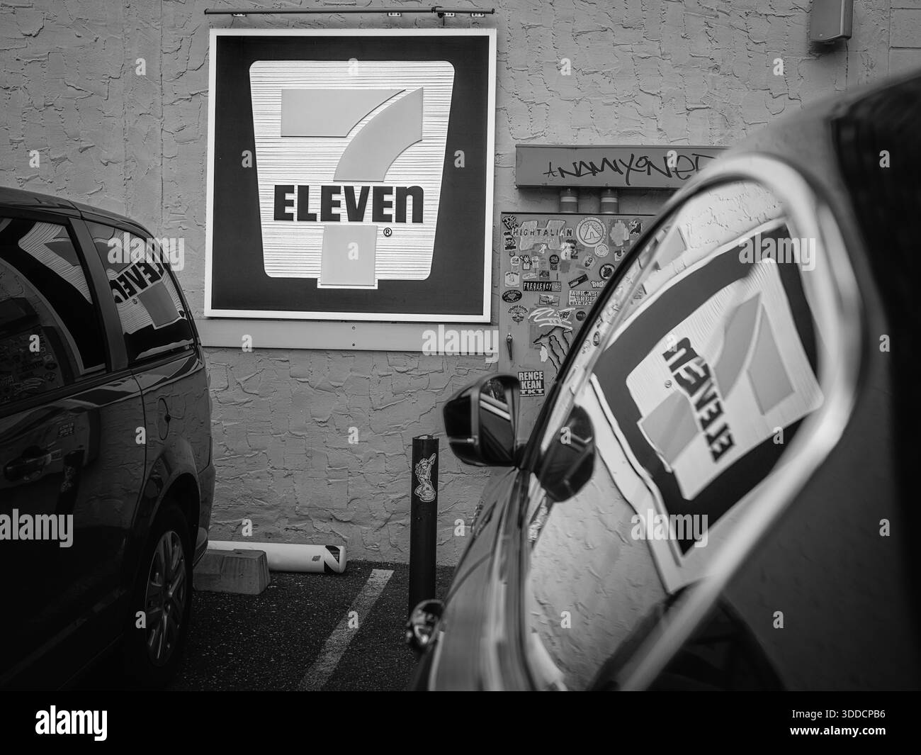 A 7-Eleven convenience store sign is photographed between parked cars in a parking area in New York, presenting a monochrome urban street scene. Stock Photo