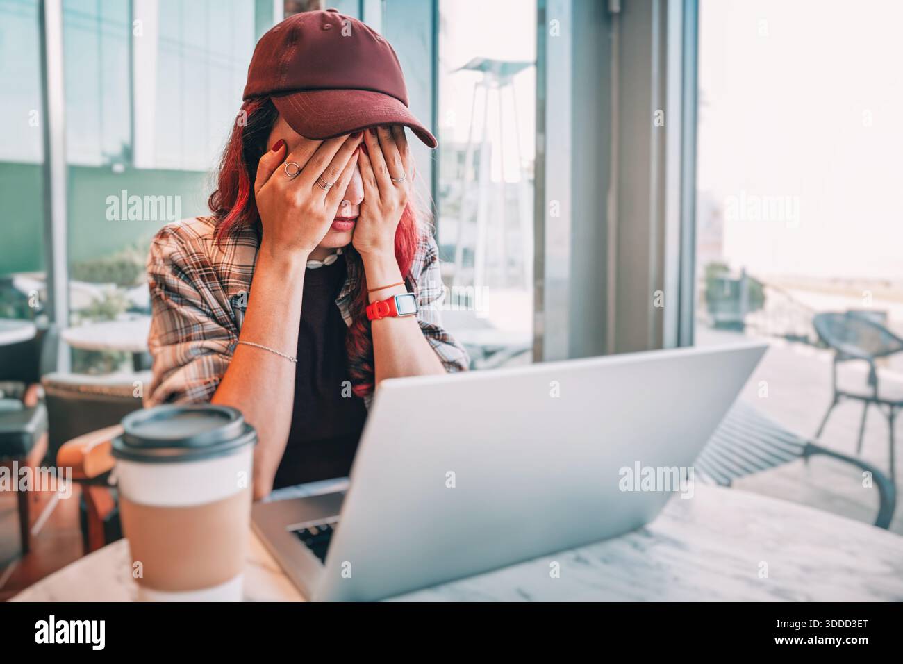 Young woman wearing a cap and plaid shirt, covering her face with hands, feeling overworked and stressed while using a laptop Stock Photo